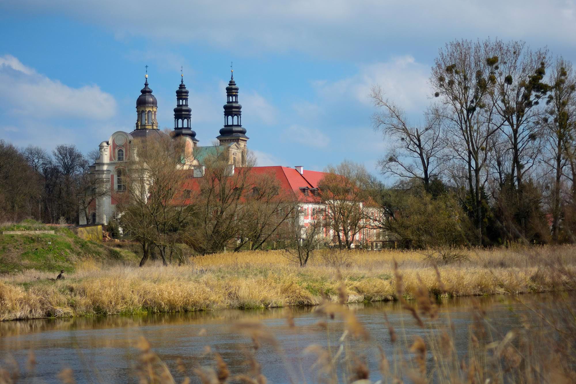 barokowy klasztor z trzema wieżami za trzcinami nad rzeką, czerwony dach i nagie drzewa wczesną wiosną