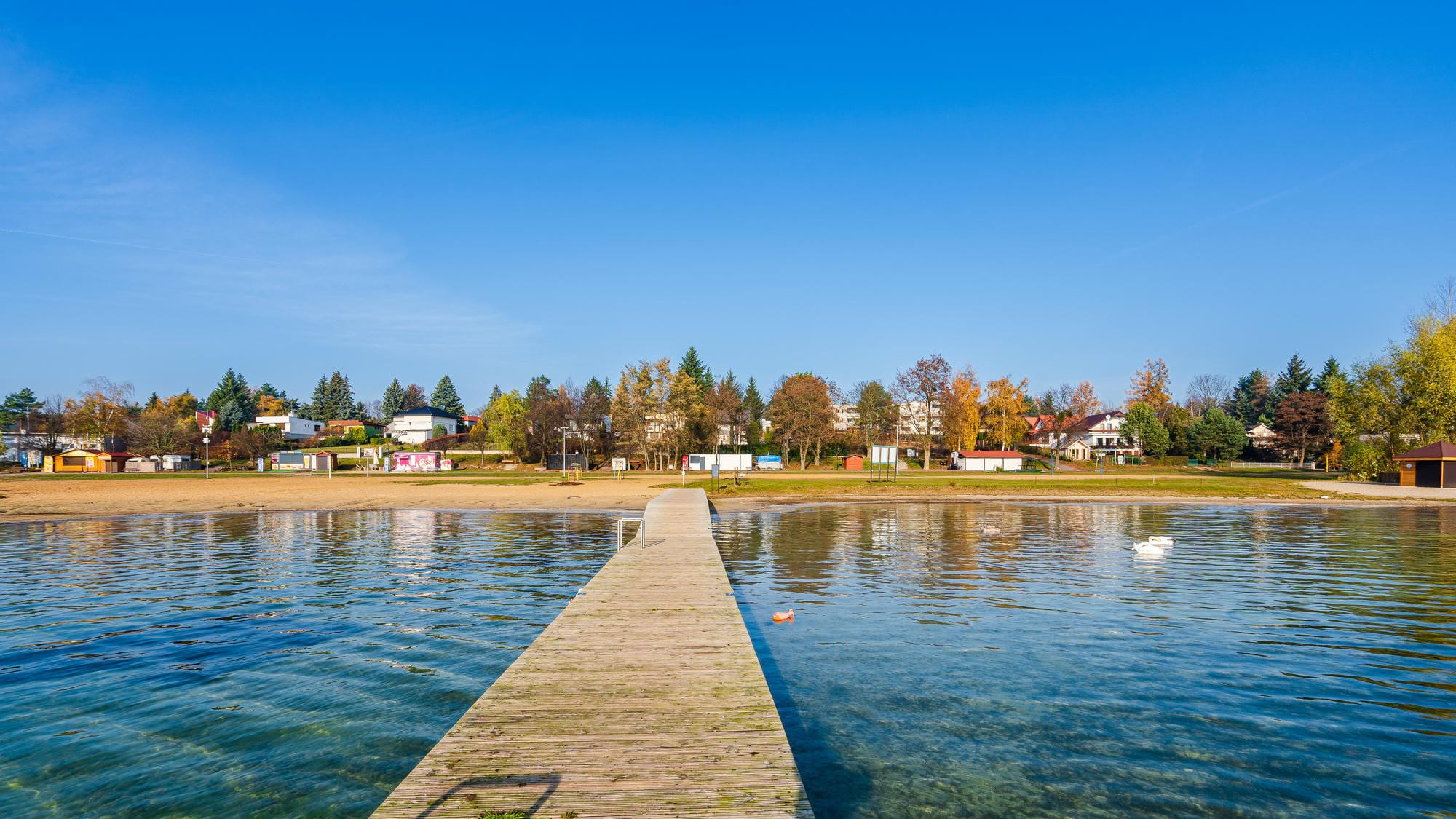 drewniany pomost prowadzący przez spokojne, przejrzyste jezioro ku piaszczystej plaży i zabudowaniom przybrzeżnym