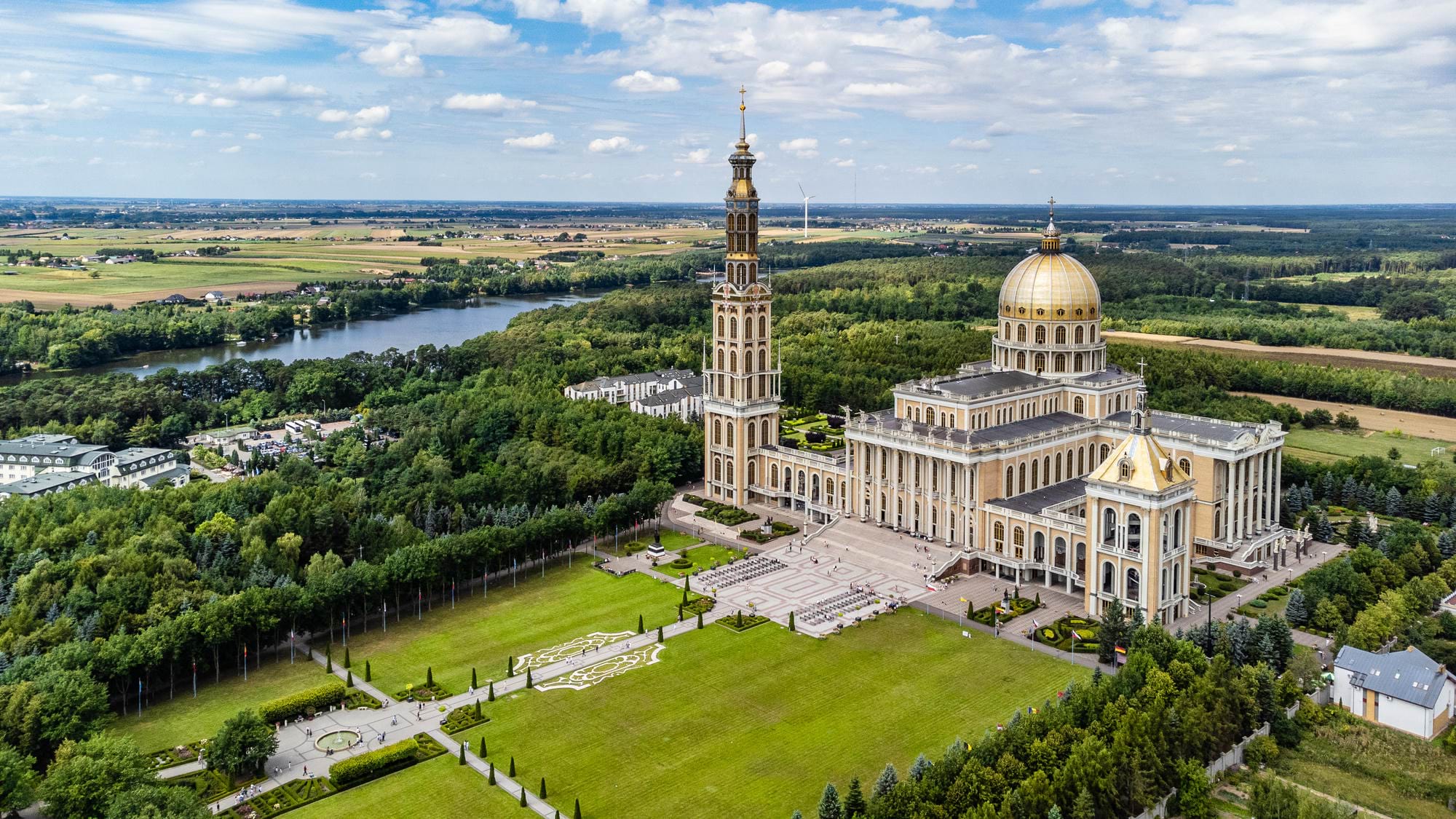 Wielka bazylika z wysoką dzwonnicą i złotą kopułą otoczona parkiem, lasem i stawem w tle